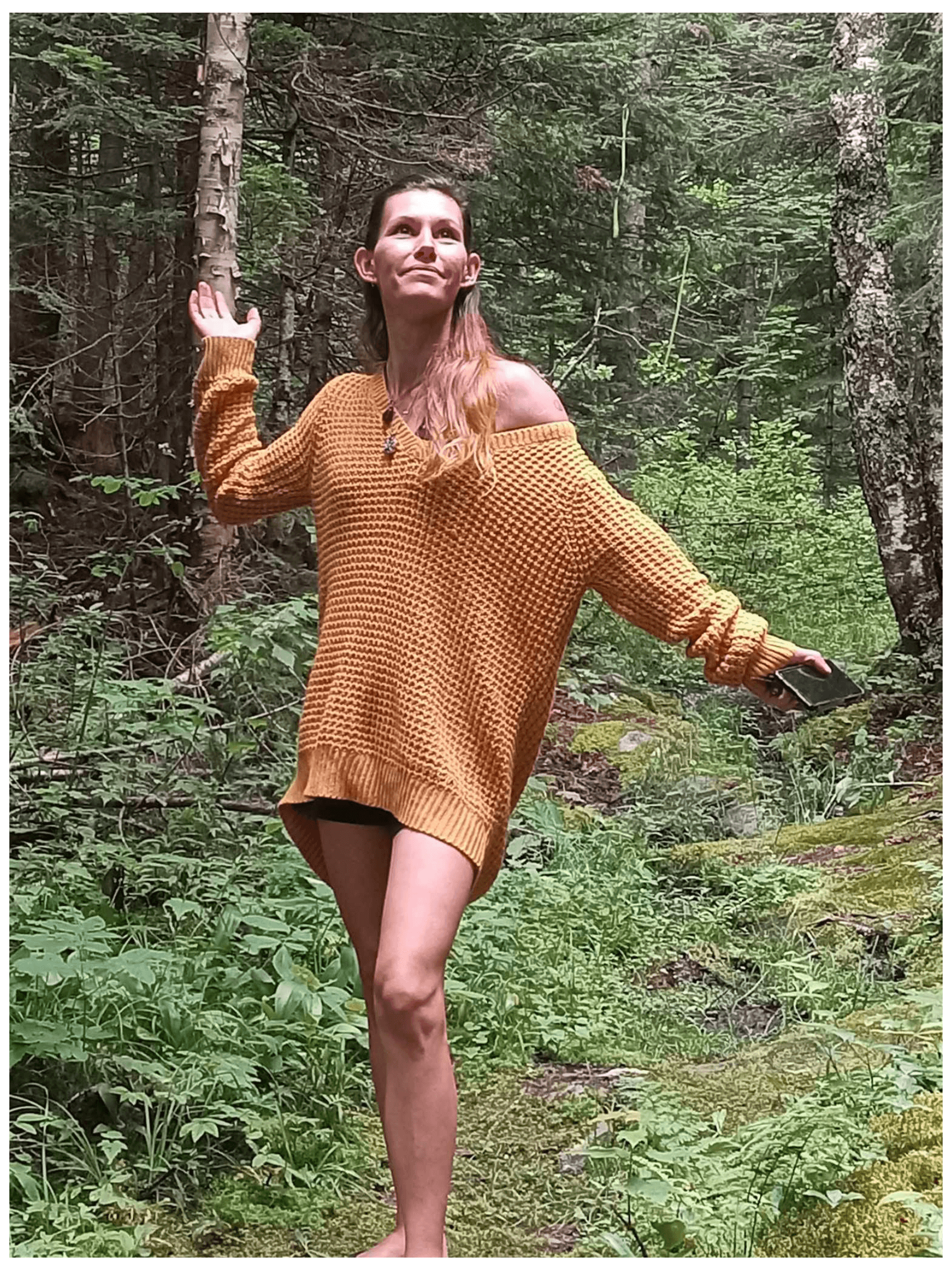 Woman in an oversized yellow sweater poses with arms outstretched in a dense green forest.